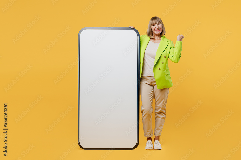 Full body elderly woman 50s year old wear green jacket white t-shirt big huge blank screen mobile cell phone smartphone with area do winner gesture isolated on plain yellow background studio portrait