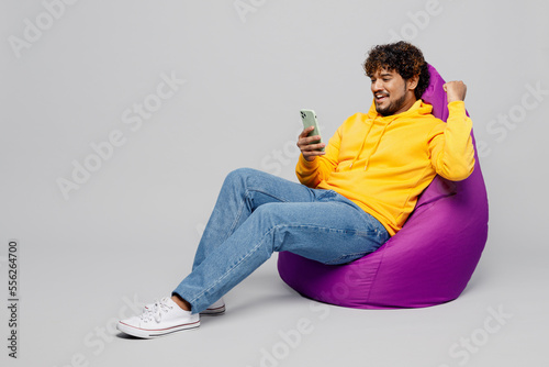 Full body young Indian man wear casual yellow hoody sit in bag chair hold in hand use mobile cell phone do winner gesture isolated on plain grey background studio portrait. People lifestyle portrait.