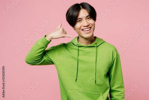 Young smiling man of Asian ethnicity wear green hoody look camera doing phone gesture like says call me back isolated on plain pastel light pink background studio portrait. People lifestyle concept. © ViDi Studio