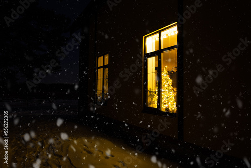 Cozy festive window of the house outside with the warm light of fairy lights garlands inside - celebrate Christmas and New Year in a warm home. Christmas tree  bokeh  snow on pine trees and snowfall