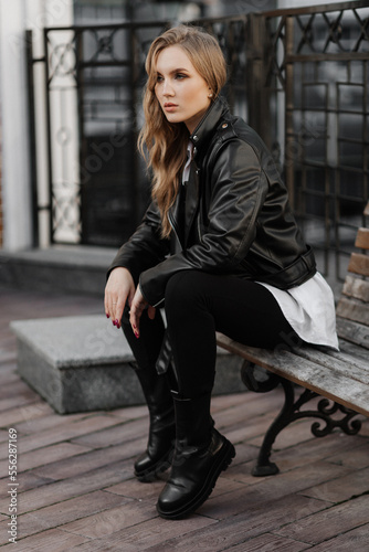 Fashionable  blonde woman model with  black leather jacket and style sunglasses sitting on a bench at the city  © Ankor_stock.