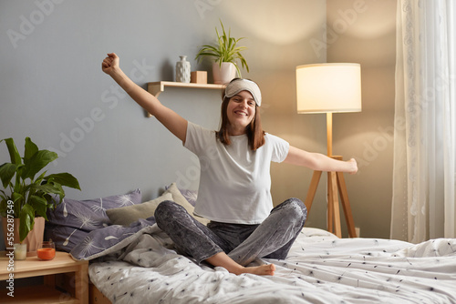 Indoor shot of smiling satisfied delighted woman wearing sleeping mask sitting on bed in cozy bedroom and stretching her arms, waking up in good mood, enjoying weekend morning.