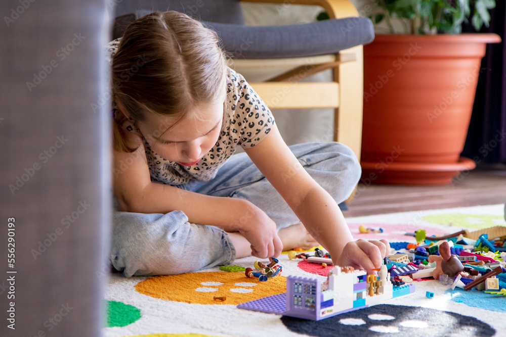 Fototapeta premium Little blonde girl plays with colorful toy blocks at home sitting on the floor in the living room. Builds and stacks. Educational games for children.
