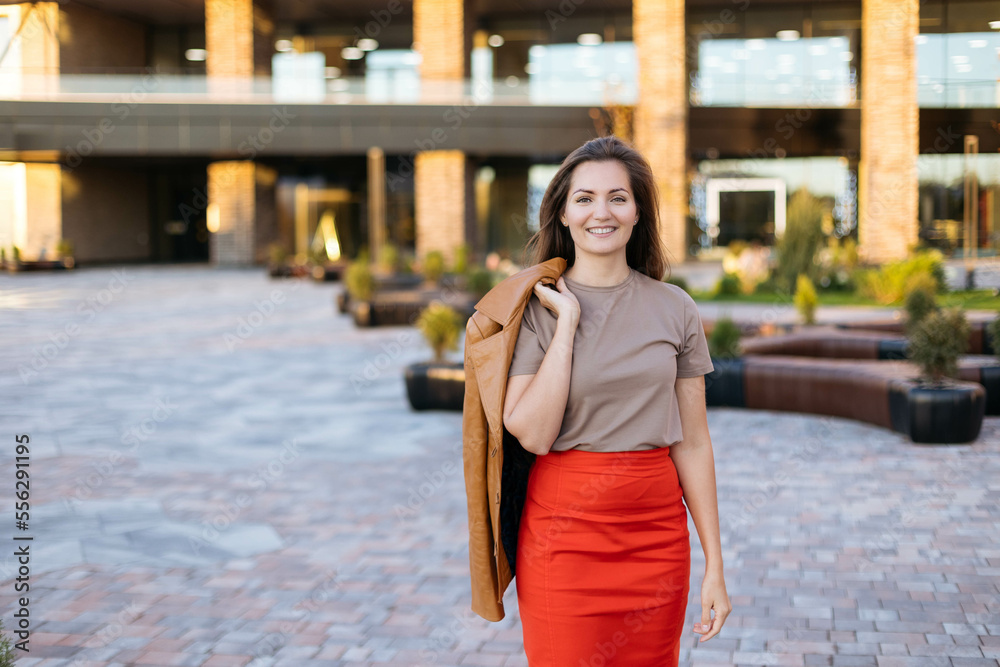 Young beautiful woman walks through the city at sunset, holding an orange jacket in her hands, looks at the camera