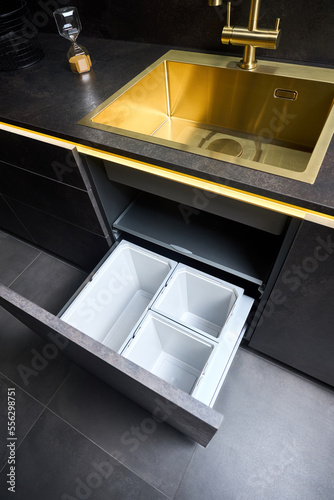 Kitchen waste bin cabinet. Pull out kitchen drawer for waste bin separate waste collection under golden sink basin and faucet black glass stoneware toptable contemporary flat design. photo