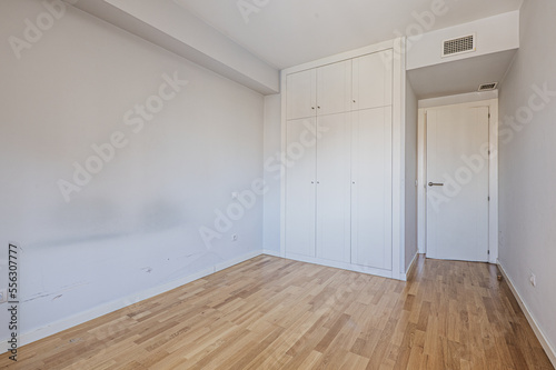 Empty room with blue walls, a three-section built-in wardrobe with mezzanines with smooth white doors, air conditioning through ducts in the ceiling and oak wood floors.