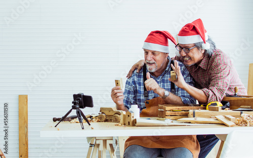 Two senior old retired men wearing Santa hat  smiling  recording live streaming video clip to social media for selling DIY wooden figure products as hobby  celebrating New year  Christmas Party.