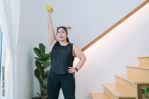 fat woman standing lifting dumbbell workout in living room. chubby woman raising arm using dumbbell building muscle. motivated overweight woman workout holding exercise tool lifting dumbbell up down photo