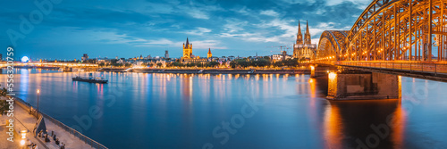 Panorama of illuminated bridge over Rhine river whith trains and tourists passing by and the Cologne Cathedral in the evening at the blue hour. photo