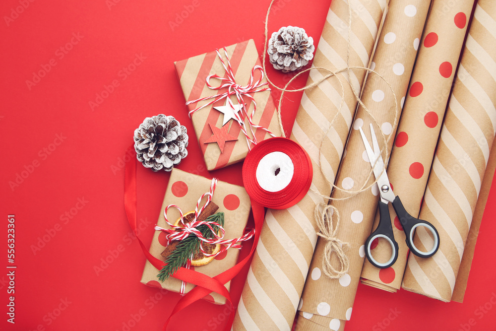 Rolls of craft papers with gift boxes, scissors and ribbon on red background