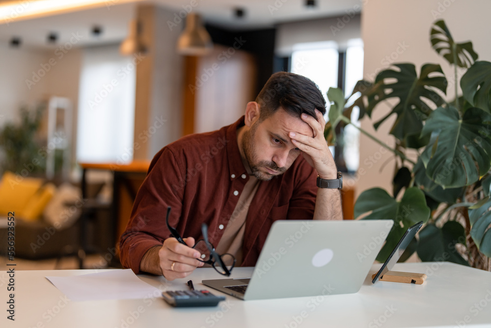 Contemplating businessman in the home office feeling sad. Tired brown haired man taking glasses off working too long at computer. Exhausted male suffer from headache sitting at desk at home.