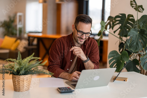 Young satisfied businessman using a laptop, typing, sitting at the desk at home. Home office. Working from home. He feels satisfied and proud of his work.