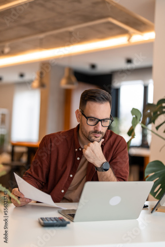 Front view of serious millennial man using laptop sitting at the working places, focused guy communicating online, writing emails, distantly working or studying on computer.