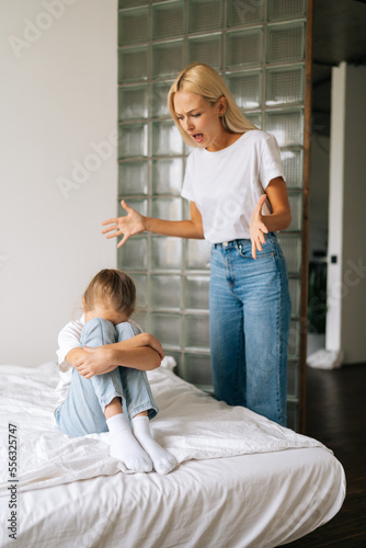 Vertical shot of sad crying little girl covering face with palm while angry young mother scolding  screaming and gesturing with hands at stubborn difficult little child daughter for bad behavior.