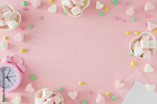 Sweet Valentines day concept. Flat lay photo of heart shaped saucers with candies and heart marshmallow, paper envelope, alarm clock on pastel pink background with copy space in the middle. © Goncharuk film