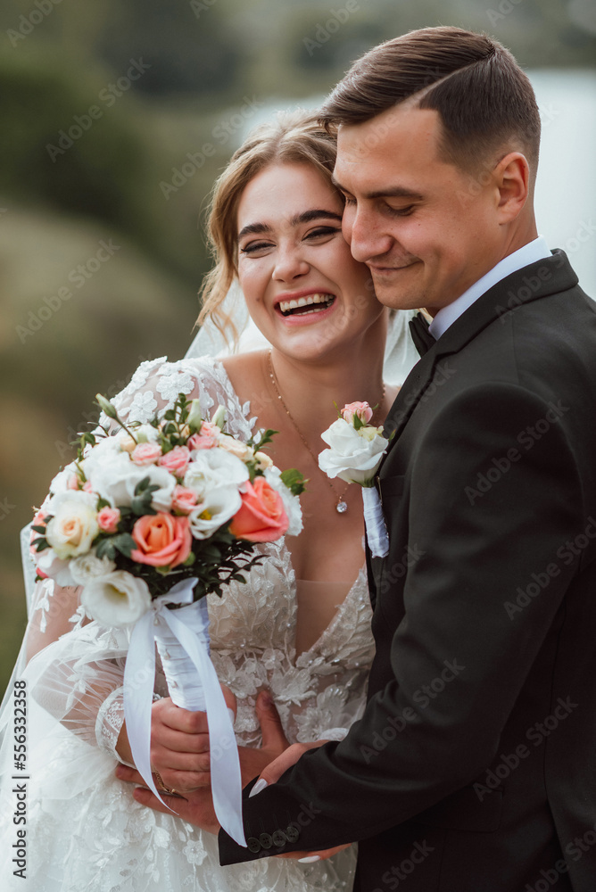 newlyweds in love on their wedding day pose for a photographer against the background of nature; he is in a dark classic jacket and she is in a white dress; a very happy and cheerful couple of people newlyweds in love on their wedding day pose for a photographer against the background of nature; he is in a dark classic jacket and she is in a white dress; a very happy and cheerful couple of people