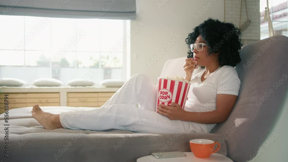 African American female watching entertaining movie on TV. Female really scared by thriller movie plot scenario and special effects. Attractive woman sitting on couch sofa with popcorn basket at home