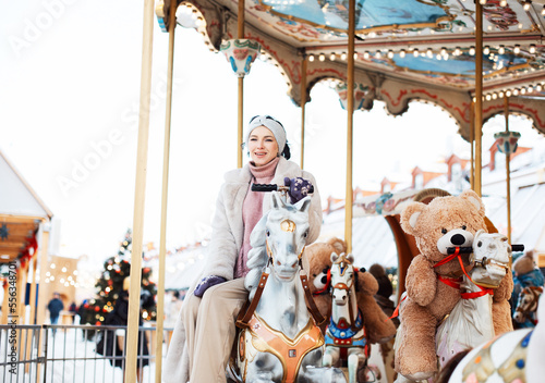 Cheerful young woman in a faux fur coat and mittens rides a carousel and has fun