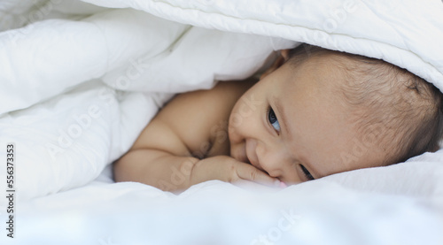 Cute adorable baby boy hidden in white blanket on the bed, laying prone and looking to the side while smiling.