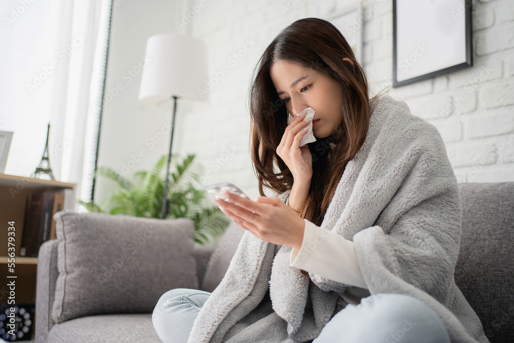 Sick asian woman sit under blanket on sofa and sneeze with tissue paper at home. She is reading check drug medicine capsule at home, suffer from flu. Cold and fever concept.