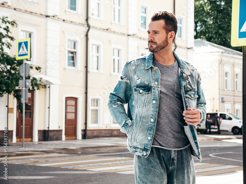 Portrait of handsome confident stylish hipster model. Sexy man dressed in jeans jacket. Fashion male posing in the street. In Europe city. Outdoors at sunny day