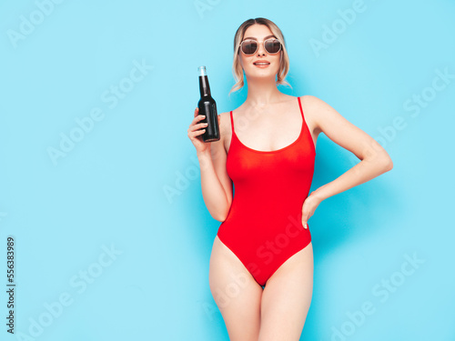 Young blond model in summer red swimwear bathing suit. Sexy carefree woman having fun and going crazy. Female posing near blue wall in studio. Drinking fresh cocktail smoozy drink from glass bottle