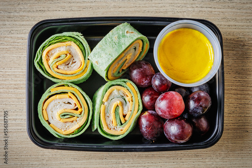 Spinach tortilla pinwheel consisting of smoked turkey, cheddar cheese, and fresh spinach. Includes grapes and sauce in a to-go container.