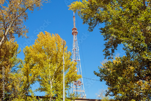 Huge city television tower antenna and autumn yellow leaves trees on blue sky. View from below