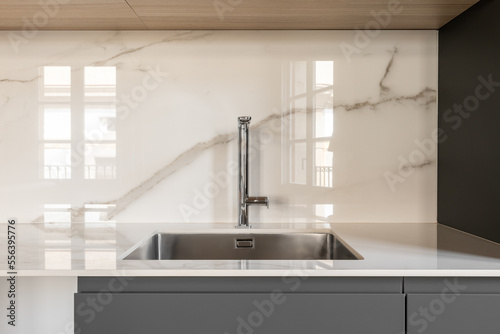 Modern small square sink in the kitchen. Fashionable built-in stainless steel sink. Interior kitchen in white and gray colors with marble tiles on the wall. White countertop and gray kitchen cabinet.