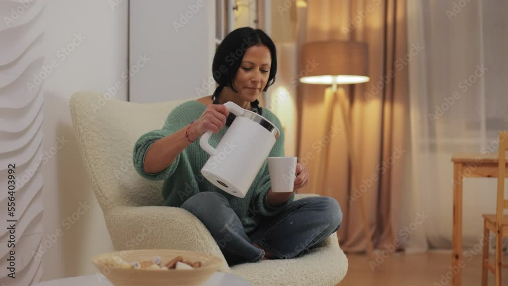Smiling middle-aged woman relaxing on a chair at home, pouring hot water from an electric kettle into a cup of tea, taking a cookie or candy from a plate and eating and drinking tea Smiling middle-aged woman relaxing on a chair at home, pouring hot water from an electric kettle into a cup of tea, taking a cookie or candy from a plate and eating and drinking tea