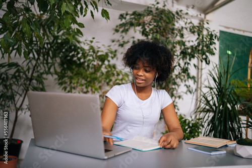 Positive curly African-American young woman in headphones writing notes, watching webinar, studying online using laptop, sitting at table in green home office room with modern biophilia design.