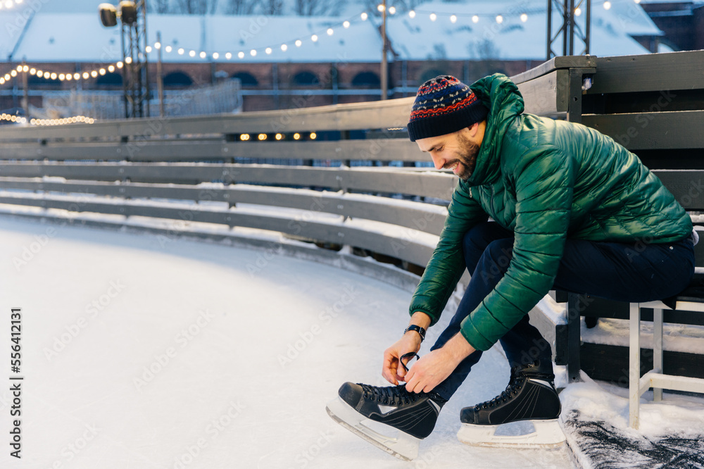 Photo of skilled male model with happy expression laces up skates, dressed in green anorak, sits on ice ring, going to be involved in his favourite hobby. Man has fun and entertainment outdoor