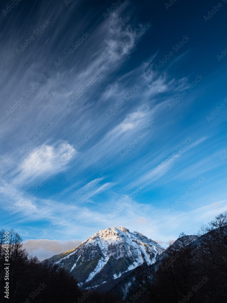 青い青空一面に白い筋雲、谷間の奥には雪を被った山、山頂に日差しが当たる。 青い青空一面に白い筋雲、谷間の奥には雪を被った山、山頂に日差しが当たる。