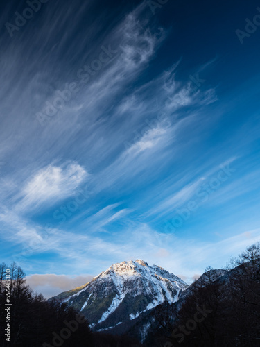 青い青空一面に白い筋雲、谷間の奥には雪を被った山、山頂に日差しが当たる。