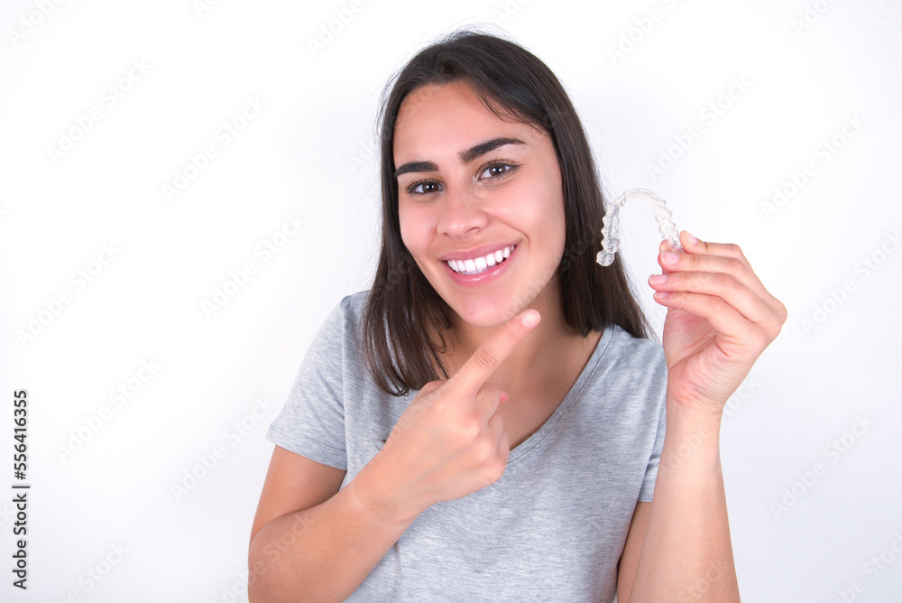Young brunette woman wearing casual clothes standing over white studio background holding an invisible aligner braces and pointing with forefinger at it. Young brunette woman wearing casual clothes standing over white studio background holding an invisible aligner braces and pointing with forefinger at it.