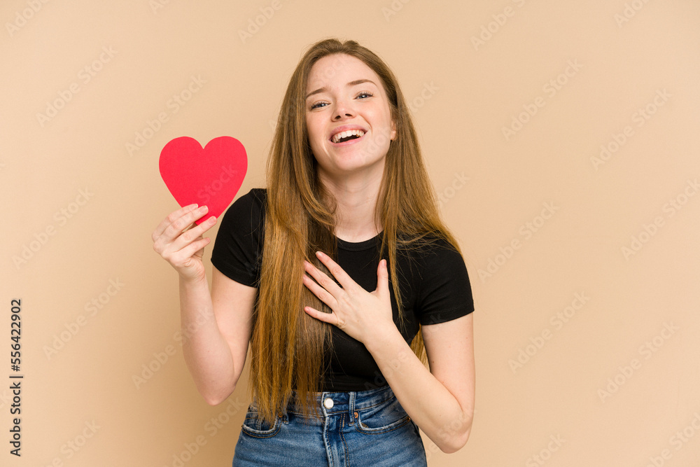 Young redhead woman holding a paper red heart cut out isolated laughs out loudly keeping hand on chest. Young redhead woman holding a paper red heart cut out isolated laughs out loudly keeping hand on chest.