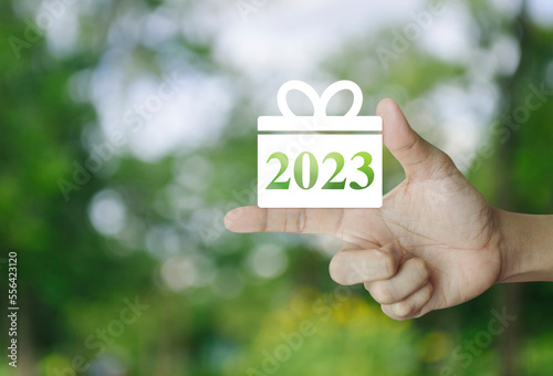 Gift box happy new year 2023 flat icon on finger over blur green tree in park, Business shopping online concept