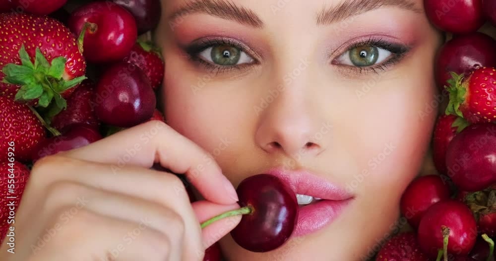 Beautiful woman holds cherry close to face. Beautiful woman with fashion makeup on vivid background from berries. Colorful image. Sweet fruits around female face with pink eye make-up. Art concept.. Beautiful woman holds cherry close to face. Beautiful woman with fashion makeup on vivid background from berries. Colorful image. Sweet fruits around female face with pink eye make-up. Art concept..