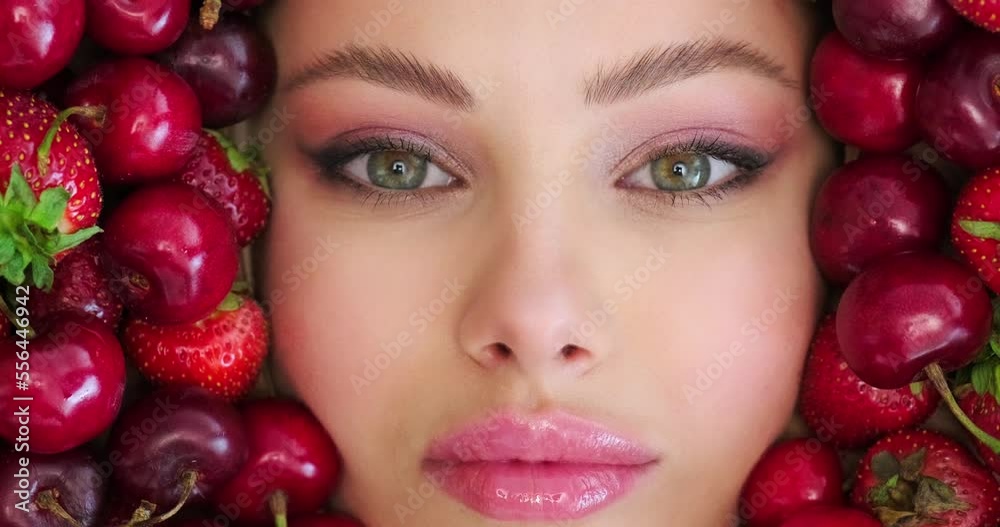 Beautiful woman holds cherry close to face. Beautiful woman with fashion makeup on vivid background from berries. Colorful image. Sweet fruits around female face with pink eye make-up. Art concept.. Beautiful woman holds cherry close to face. Beautiful woman with fashion makeup on vivid background from berries. Colorful image. Sweet fruits around female face with pink eye make-up. Art concept..