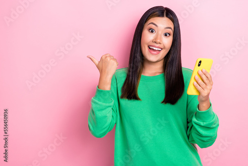 Photo of excited funky vietnamese youngster lady wear green jumper hold smartphone direct finger mockup eshop promo isolated on pink color background