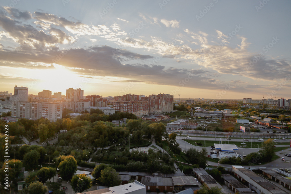 Sunset illuminates busy city with residential area surrounded by park with green trees and road. Sunlight breaks through cumulus clouds