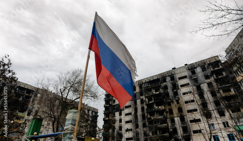 tricolor flag of Russia against the background of destroyed and burnt houses in Ukraine