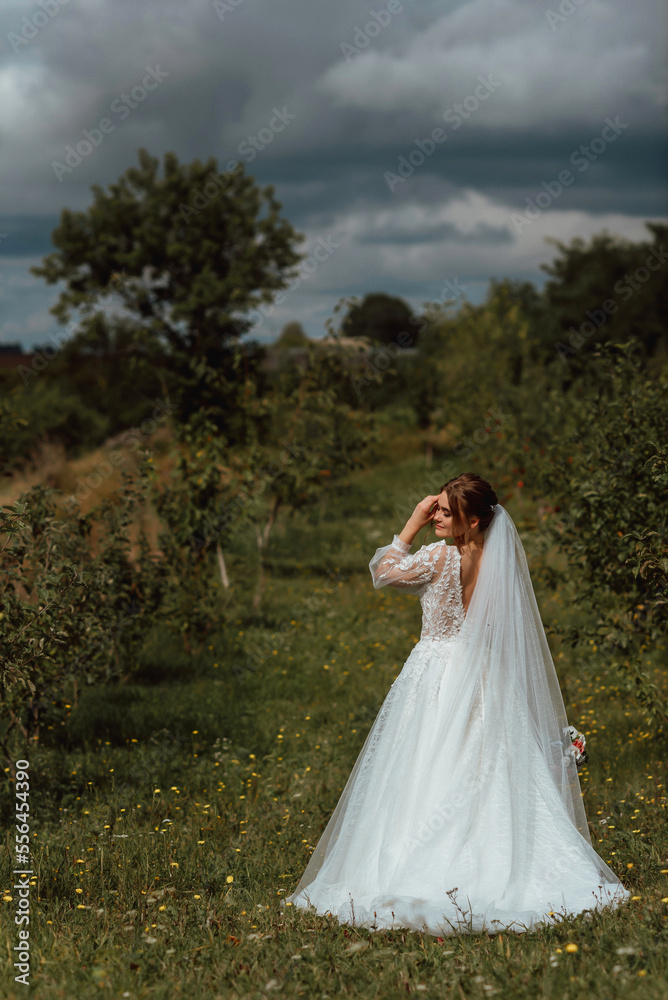 a young beautiful bride poses with a wedding bouquet against the background of nature; the girl is dressed in a white wedding dress; a moment on the wedding day; happy girl expresses her emotions