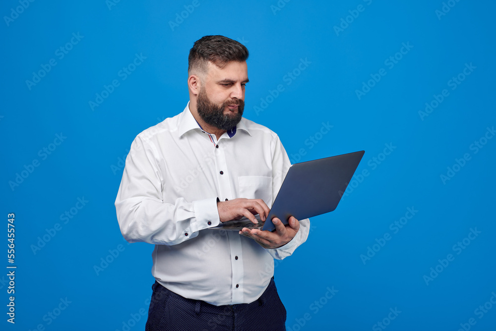 A bearded Caucasian man in a white shirt on a blue background holds a laptop. A solid man with a laptop.