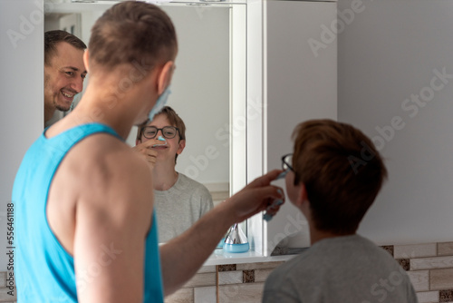 Young father and his teenage son with shaving foam on his nose. Dad and son in bathroom in front of mirror together.