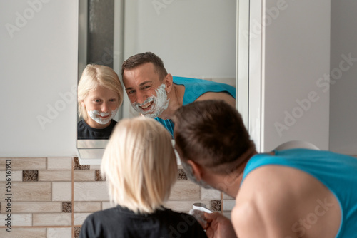 Smiling dad and little son with shaving foam on face look in bathroom mirror. Dad and son shave and having fun