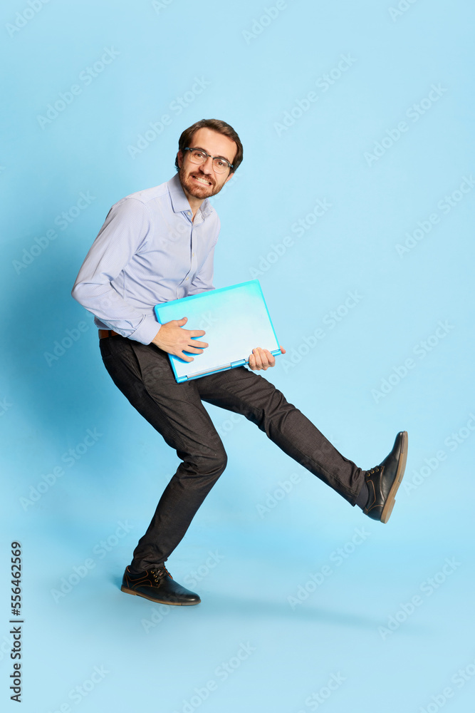 Cheerful Caucasian man, businessman in office dress code having fun, dancing with laptop over blue background. Positive emotions, work, happiness