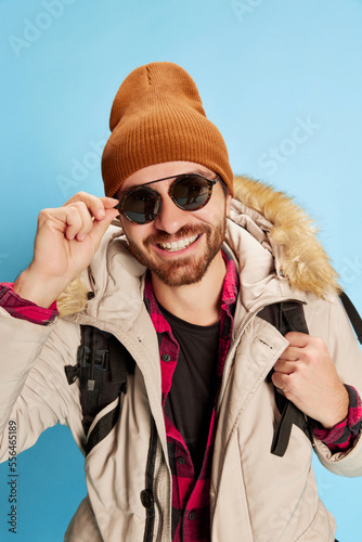 Fashionable mature man in winter coat and hat with happy facial expression looking at camera isolated on blue background. Holidays, travel, adventure, ad