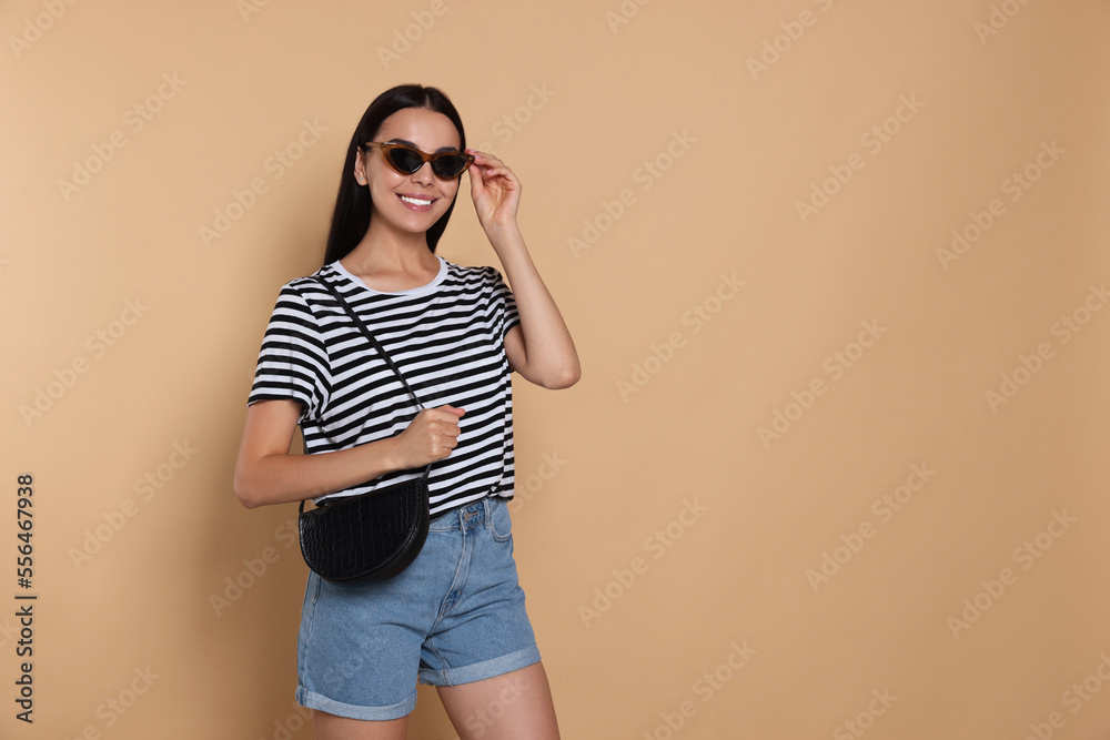 Beautiful young woman with stylish bag in sunglasses on beige background, space for text Beautiful young woman with stylish bag in sunglasses on beige background, space for text