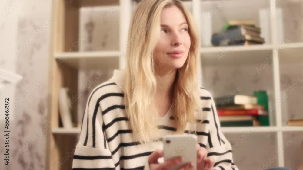 Dreamy soft shot of blond woman hold smartphone scrolling social media texting reading morning news while sitting on sofa Young female relaxing and chatting on mobile phone next to bookshelf at home Dreamy soft shot of blond woman hold smartphone scrolling social media texting reading morning news while sitting on sofa Young female relaxing and chatting on mobile phone next to bookshelf at home